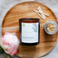 a amber glass candle on a sustainable bamboo plate with a fresh peony and matches 